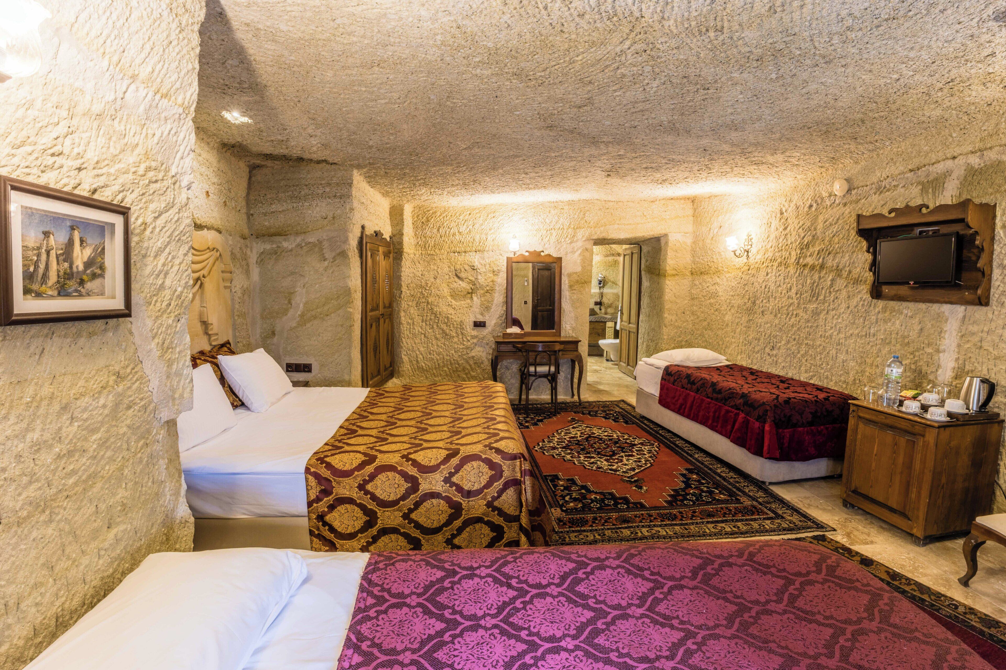 Una camera d'albergo con parete in pietra, un letto con copripiumino decorato, un secondo letto, un comodino in legno con una lampada e un televisore fissato al muro.