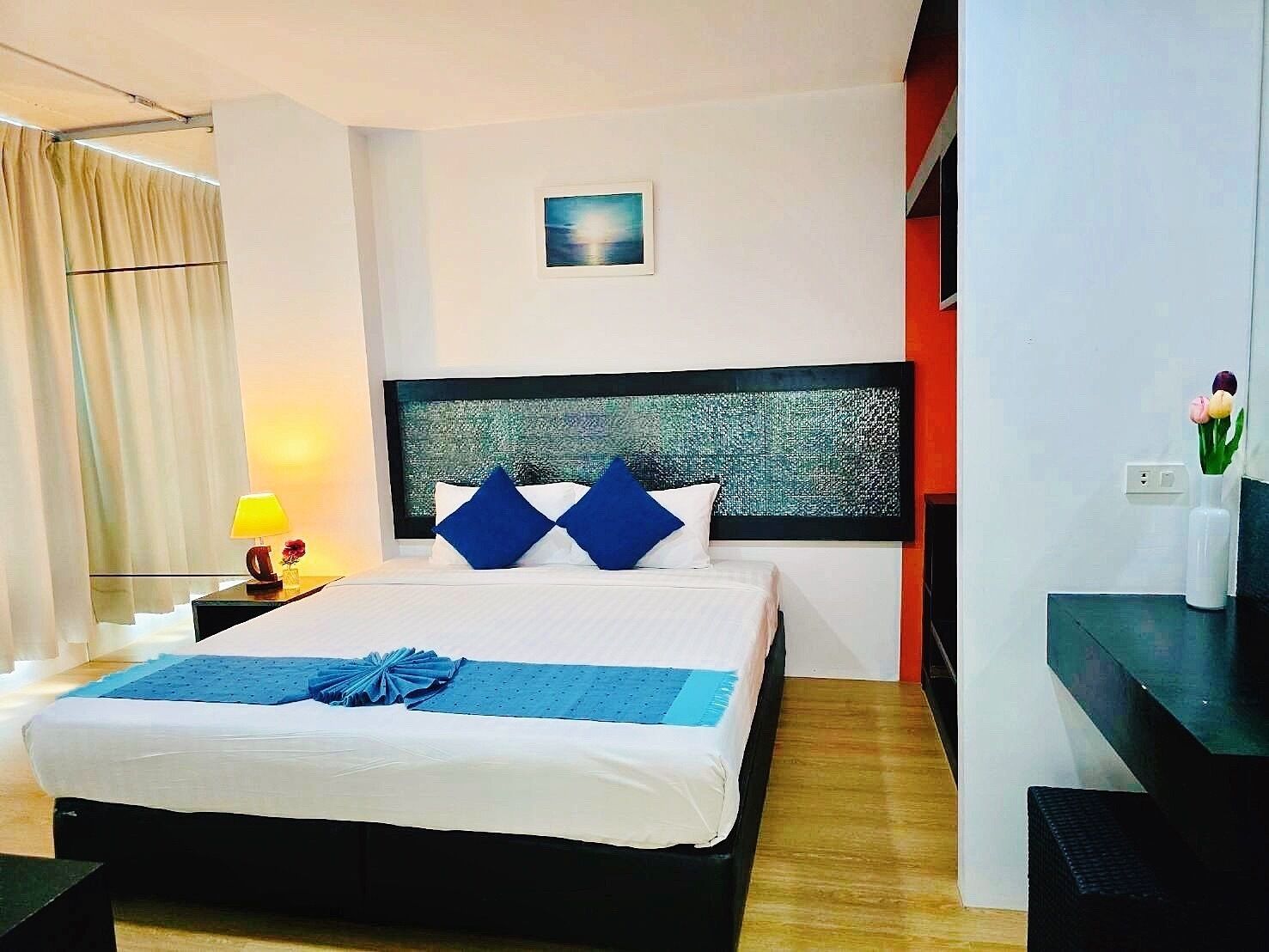 A hotel room with a large bed, a nightstand with a lamp, a window with curtains, and a small painting on the wall.