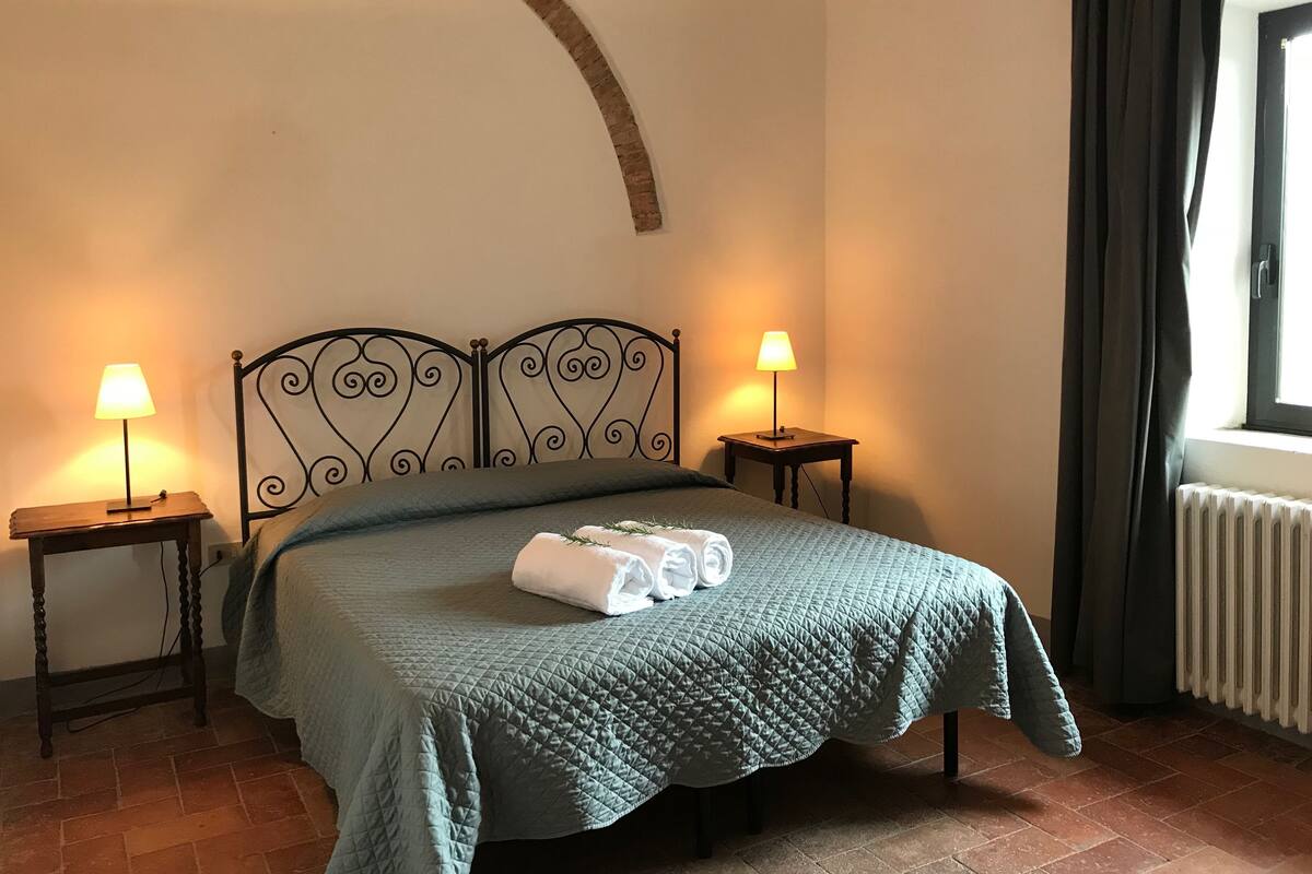 Un lit bien fait, recouvert d’une courtepointe verte matelassée, avec deux lampes de chevet et une fenêtre agrémentée de rideaux.