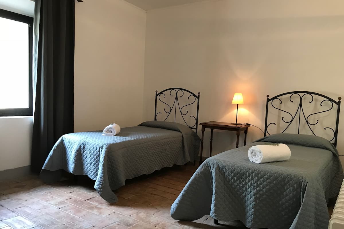 Deux lits jumeaux recouverts de couvertures vert foncé, une petite table de chevet avec une lampe et une fenêtre avec des rideaux foncés.