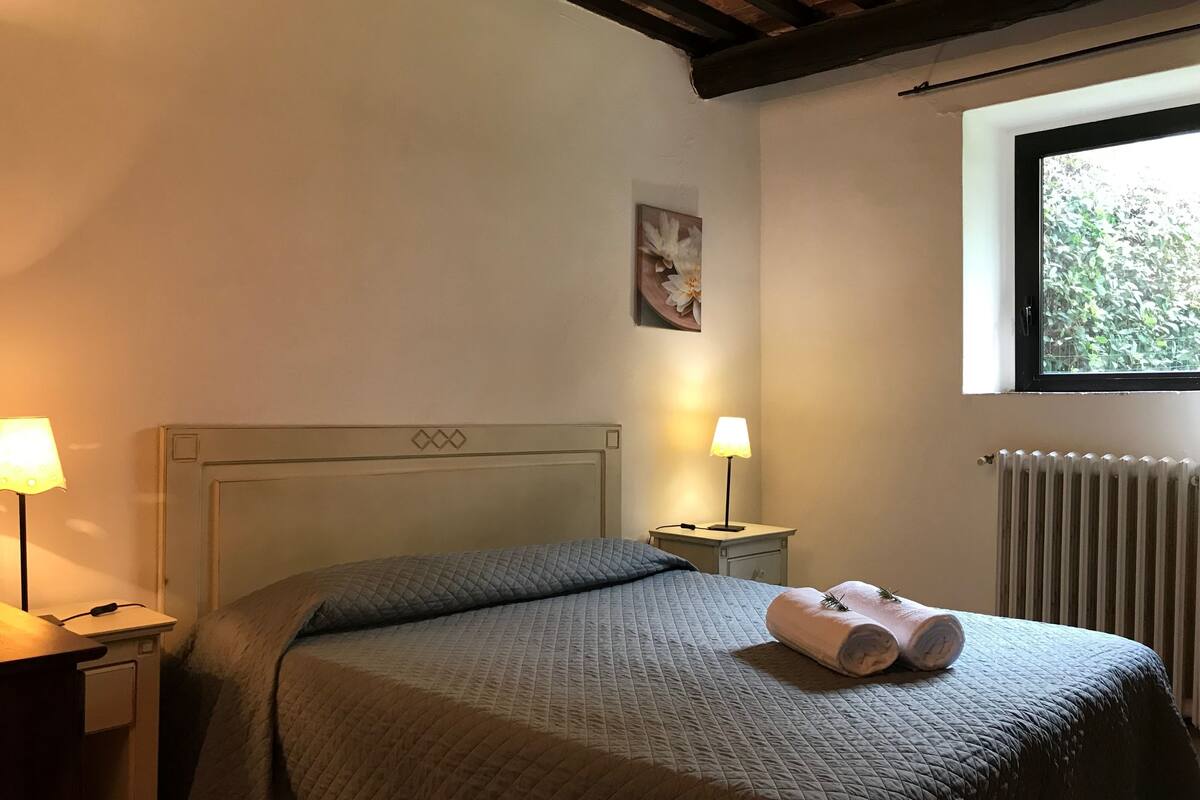 Un lit bien fait, recouvert d’une couverture matelassée, deux lampes de chevet, un radiateur et une fenêtre donnant sur un paysage verdoyant.
