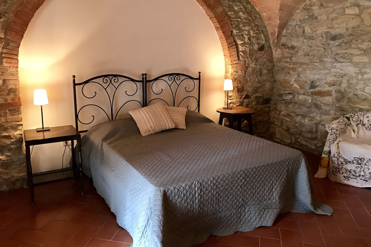 Une chambre à coucher avec une arche en pierre, un lit recouvert d’une couverture matelassée, deux lampes de chevet et une chaise agrémentée d’un coussin à motifs.