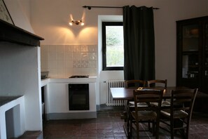 Et lille køkken med hvide fliser, indbygget ovn, et spisebord med stole og et vindue med gardiner.