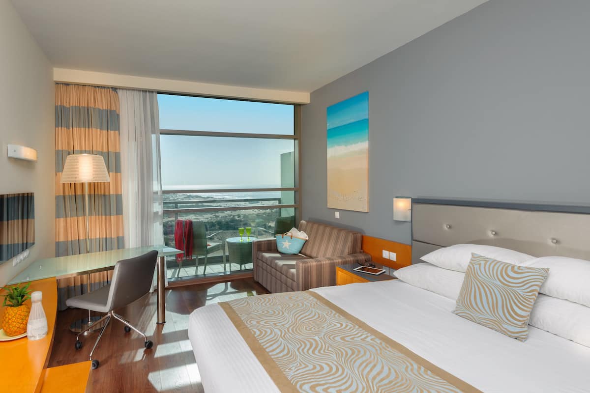 Habitación de hotel con cama, escritorio, silla, una pequeña mesa con silla y un ventanal con vistas.