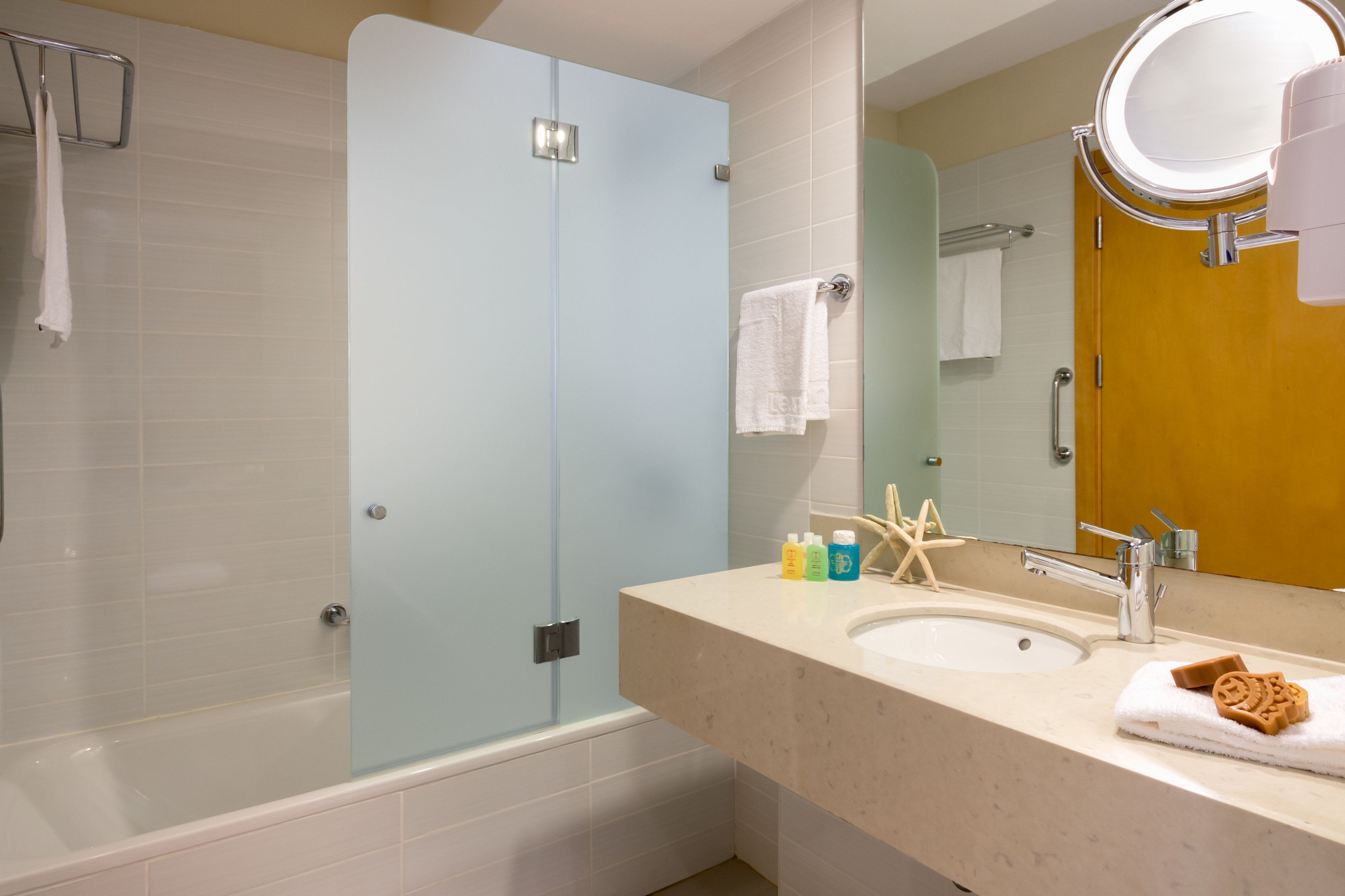 A bathroom with a glass shower enclosure, a sink with toiletries, and a mirror with a round light.