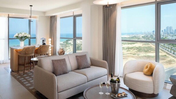 A modern hotel room with a sofa, armchair, dining area, and large windows offering a city view. - Leonardo Plaza Ashdod (Ashdod)