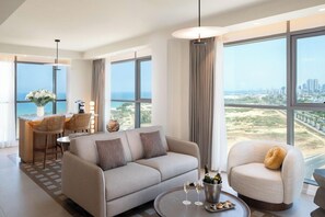 A modern hotel room with a sofa, armchair, dining area, and large windows offering a city view. - Leonardo Plaza Ashdod (Ashdod)