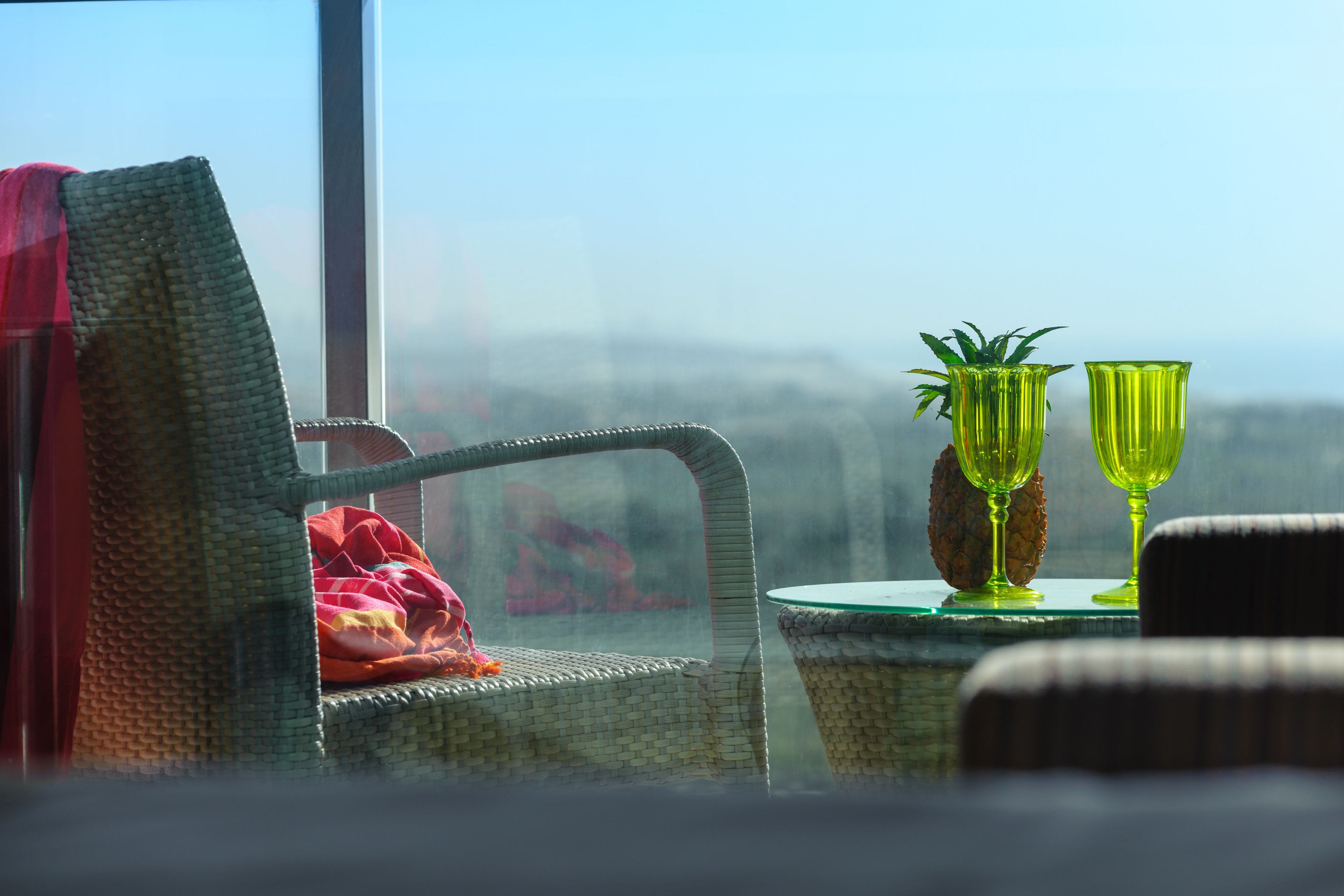 Una silla de mimbre con una manta roja, una mesita con dos jarrones de vidrio verde y una piña, y una vista de la ciudad a través de una ventana.
