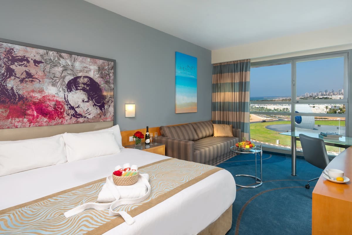 Habitación de hotel con una cama grande, un sofá, zona de comedor y vistas a la ciudad y la costa.