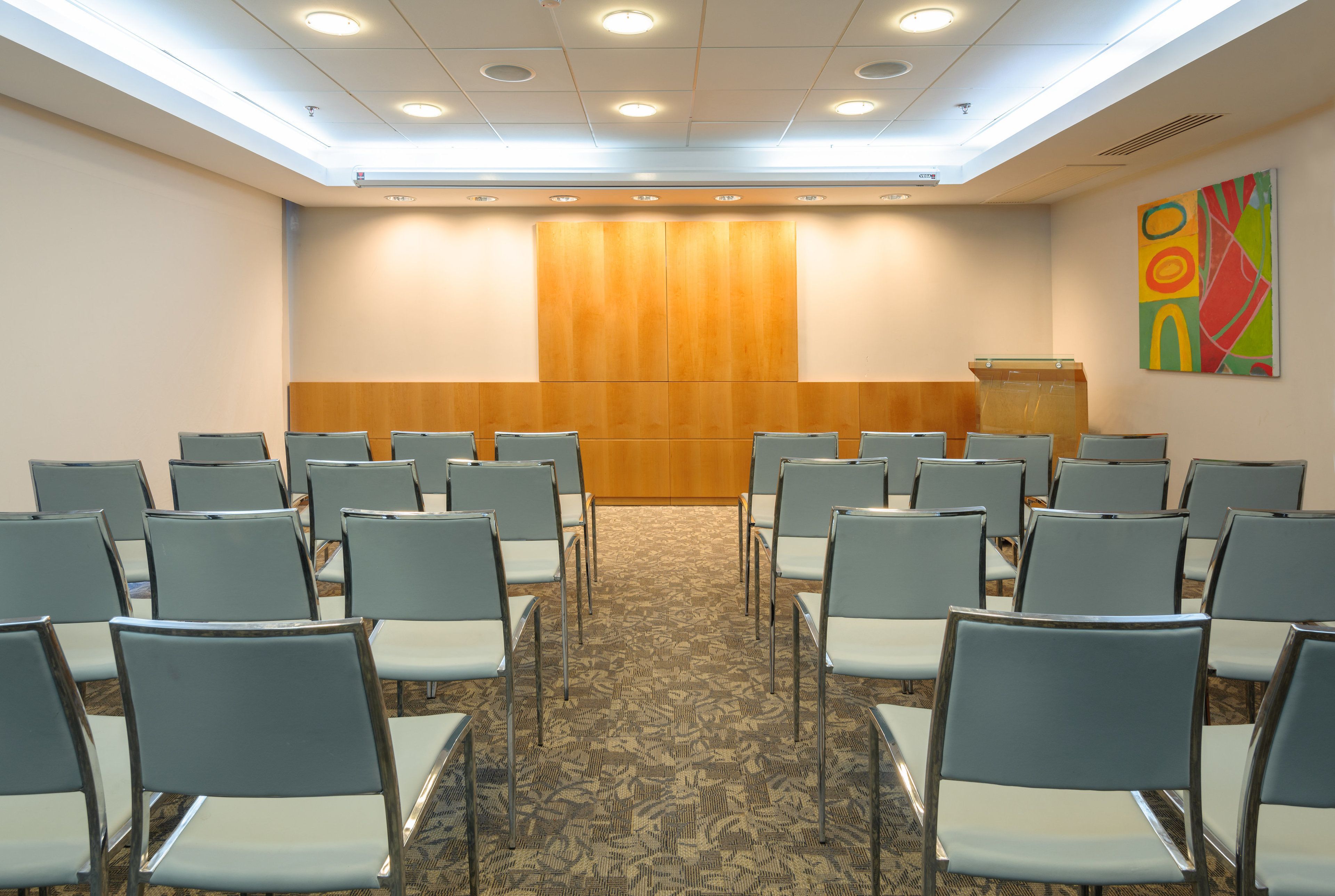 A conference room with rows of chairs facing a podium, carpeted floor, and a ceiling with recessed lighting.