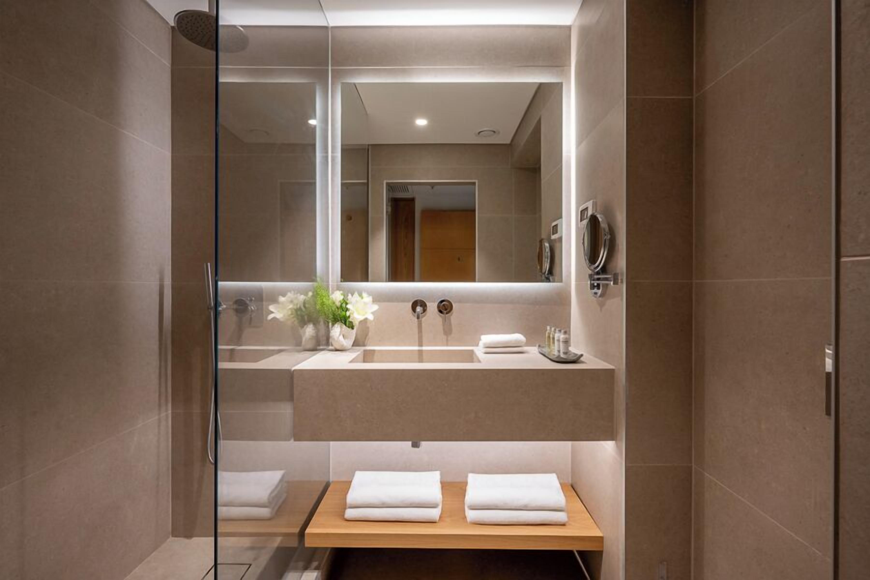 A modern bathroom with a glass shower enclosure, a large mirror, a beige sink, and a wooden vanity with towels.