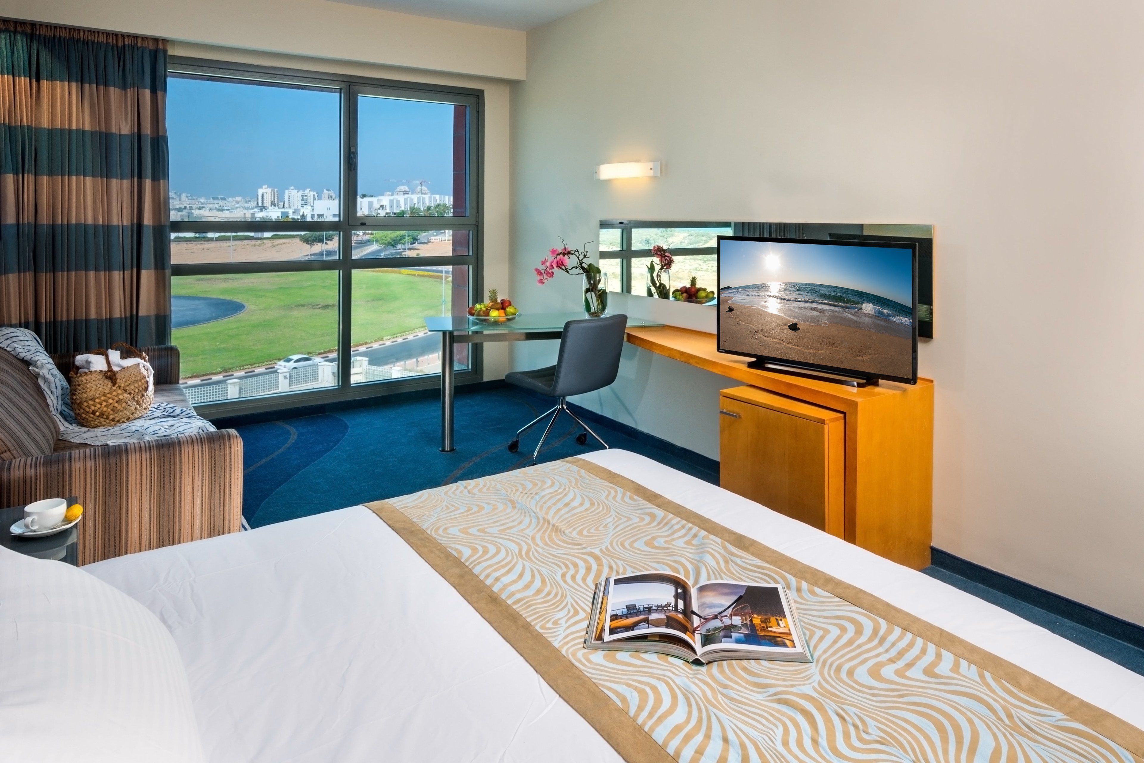 Ein Hotelzimmer mit großem Fenster, einem Bett, einem Schreibtisch mit Fernseher und Blick auf die Stadt.
