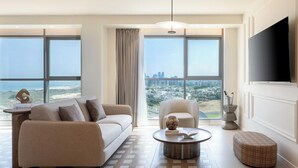 A modern living room with a sofa, armchair, coffee table, and ottoman, large windows with a city view, and a flat-screen TV mounted on the wall. - Leonardo Plaza Ashdod (Ashdod)
