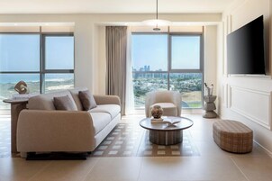 A modern living room with a sofa, armchair, coffee table, and ottoman, large windows with a city view, and a flat-screen TV mounted on the wall.