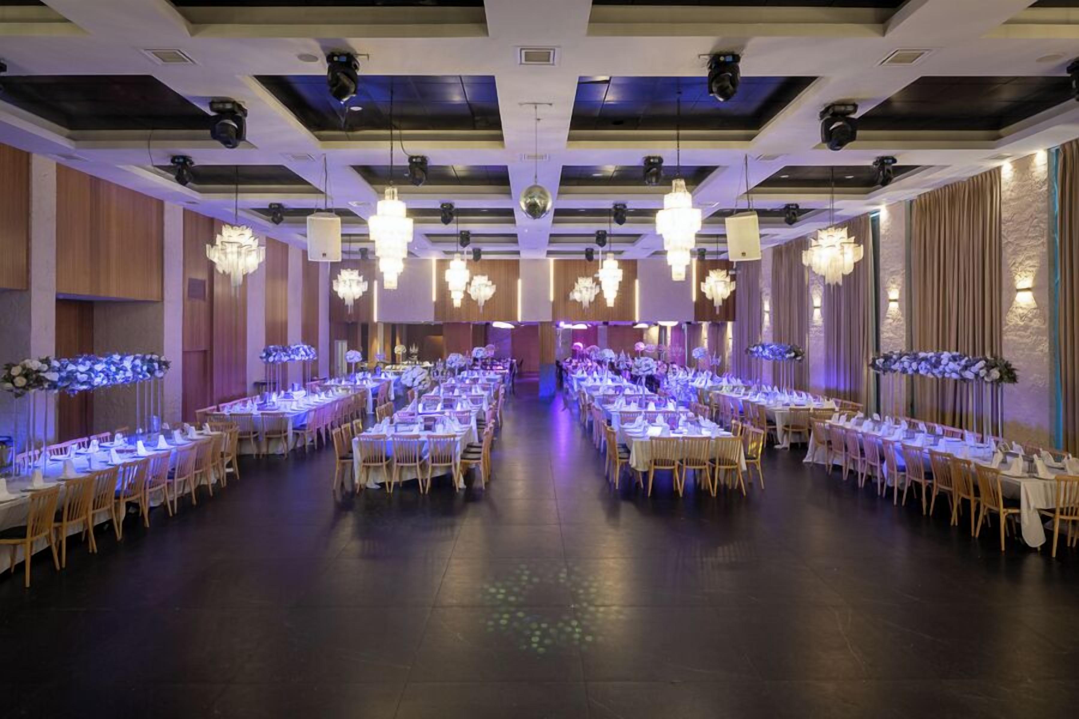 A banquet hall with multiple tables arranged in rows, chandeliers hanging from the ceiling, and a stage at the front.