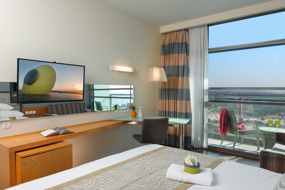 Habitación de hotel con cama, escritorio, silla, televisión, ventana con cortinas y vista a la ciudad.