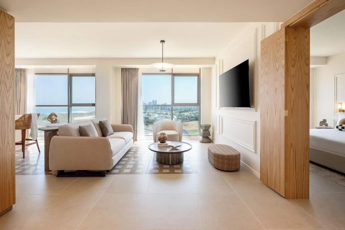 Una sala de estar moderna con sofá, mesa de centro y reposapiés, ventanas amplias con vista a la ciudad y un televisor de pantalla plana montado en la pared.