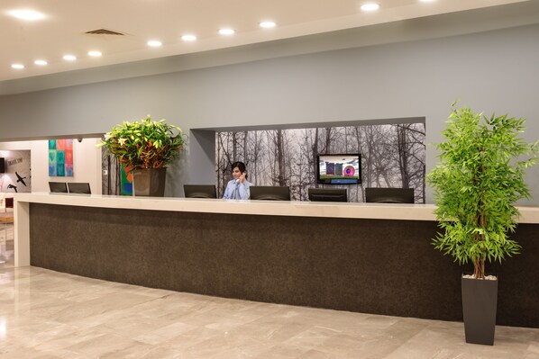Un mostrador de recepción con una recepcionista, una planta grande en maceta, una pantalla de televisión y una pared con un mural de un árbol.