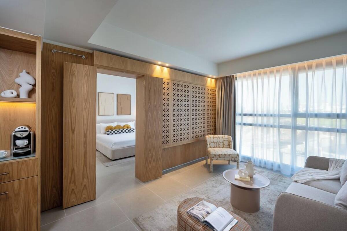 Una habitación de hotel moderna con un armario de madera, una cama, un área de descanso con sofá y sillón, una cafetera y un ventanal con cortinas.