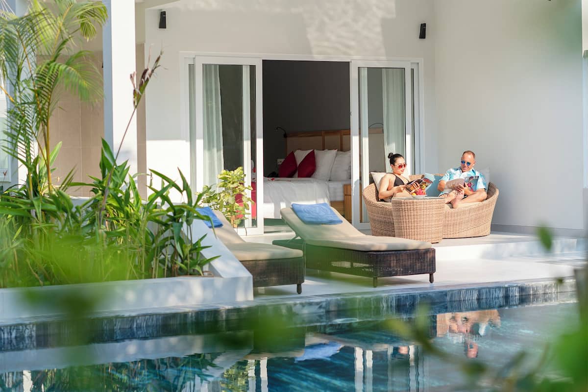 A modern hotel room with a balcony, a pool, and a bed with white linens and red pillows.