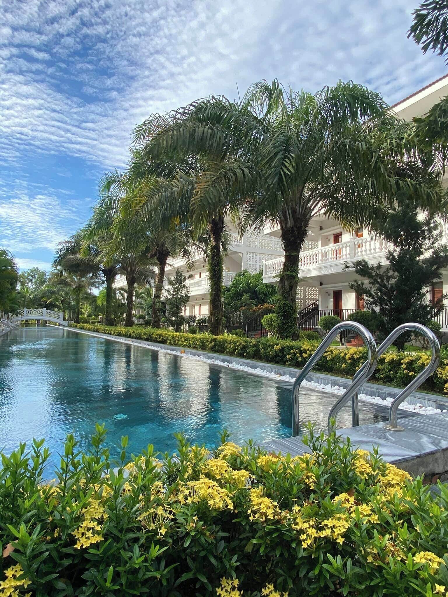 A swimming pool with a bridge, surrounded by palm trees and yellow flowers, with a building in the background.