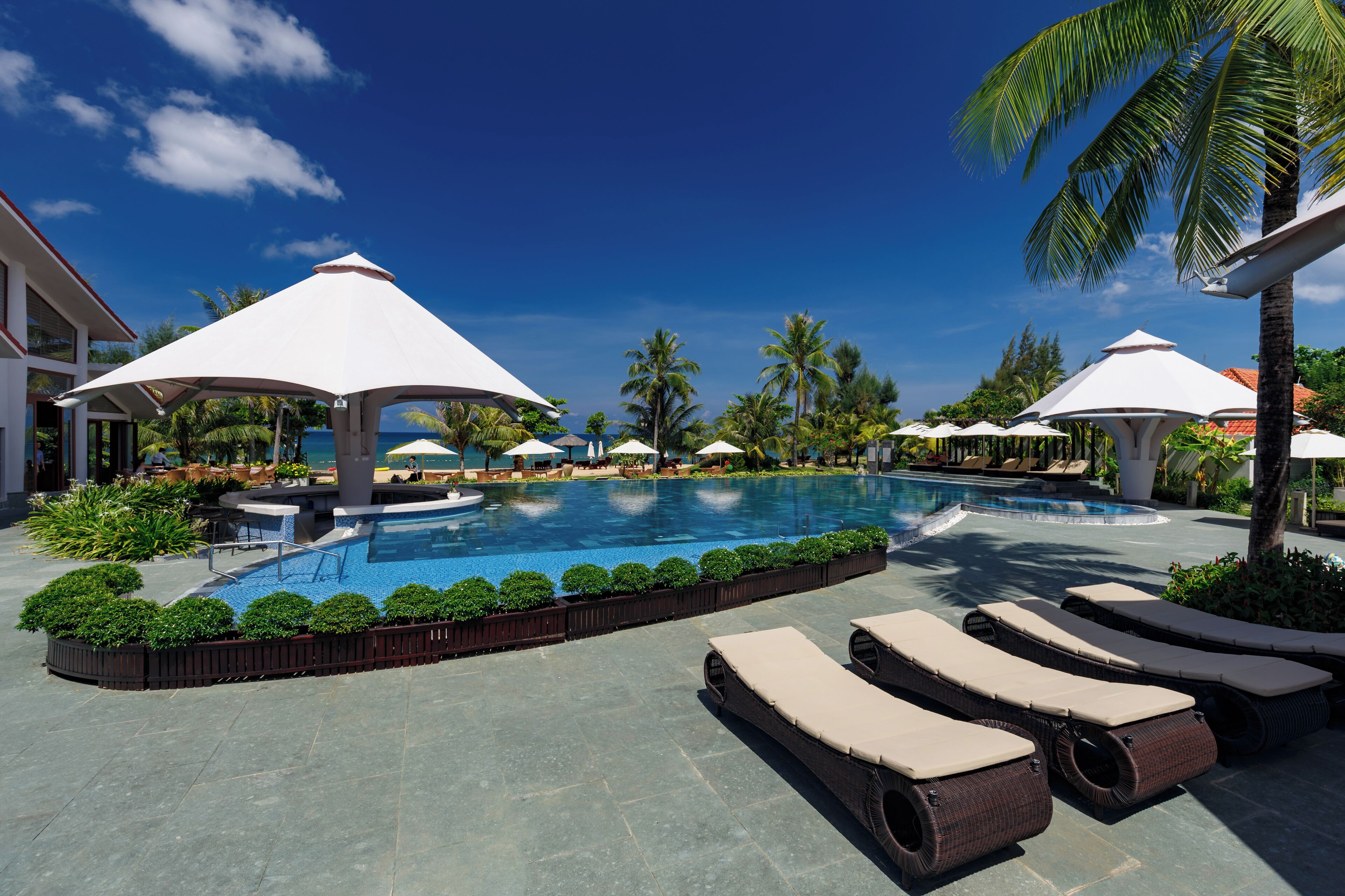 A swimming pool with lounge chairs and umbrellas, surrounded by palm trees and a clear sky.