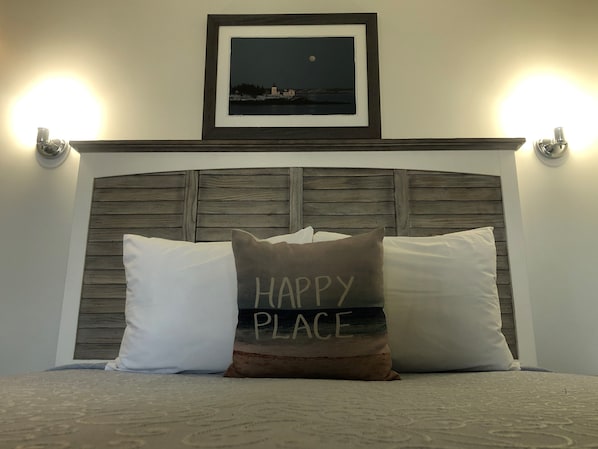 A bed with two pillows, one with "HAPPY PLACE" text, and a framed picture above the bed.