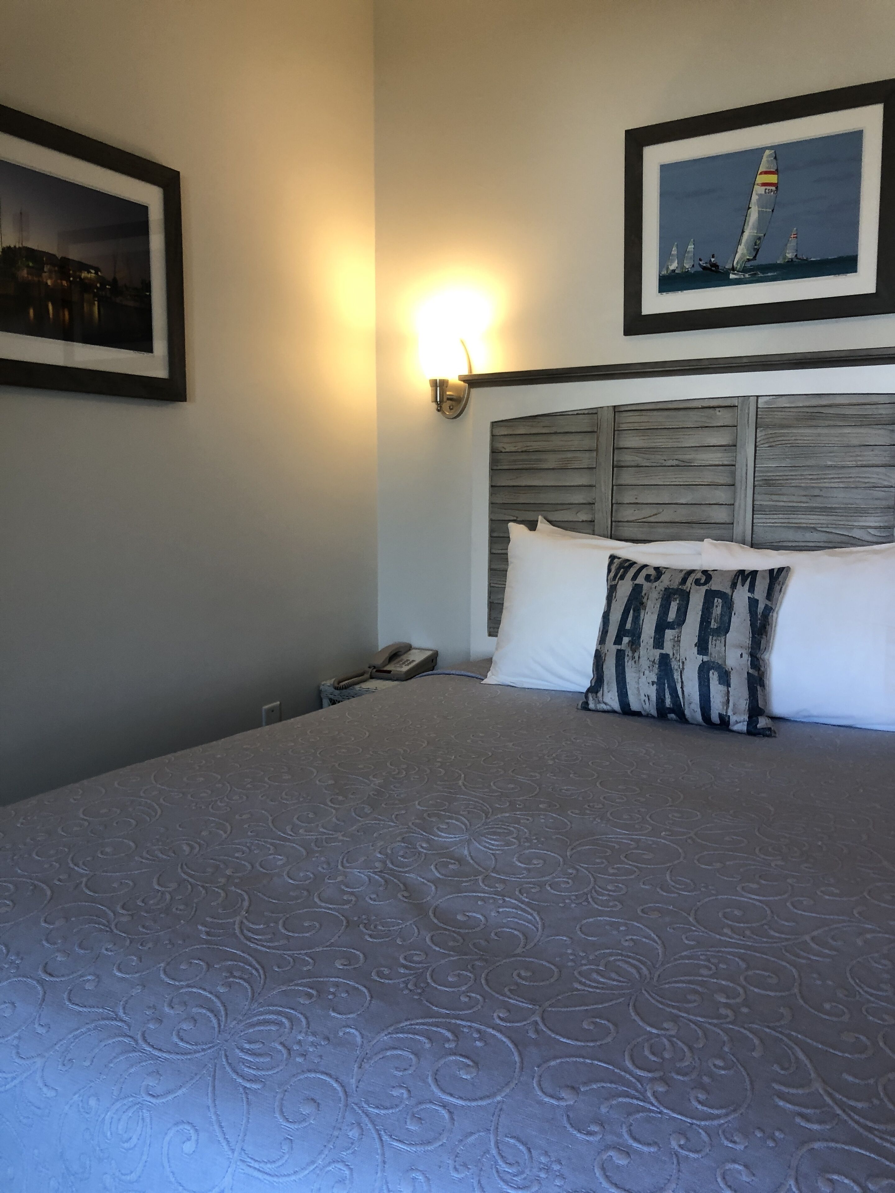 Une chambre d’hôtel avec un grand lit, une tête de lit ornée d’un panneau en bois, une lampe fixée au mur et deux tableaux encadrés.