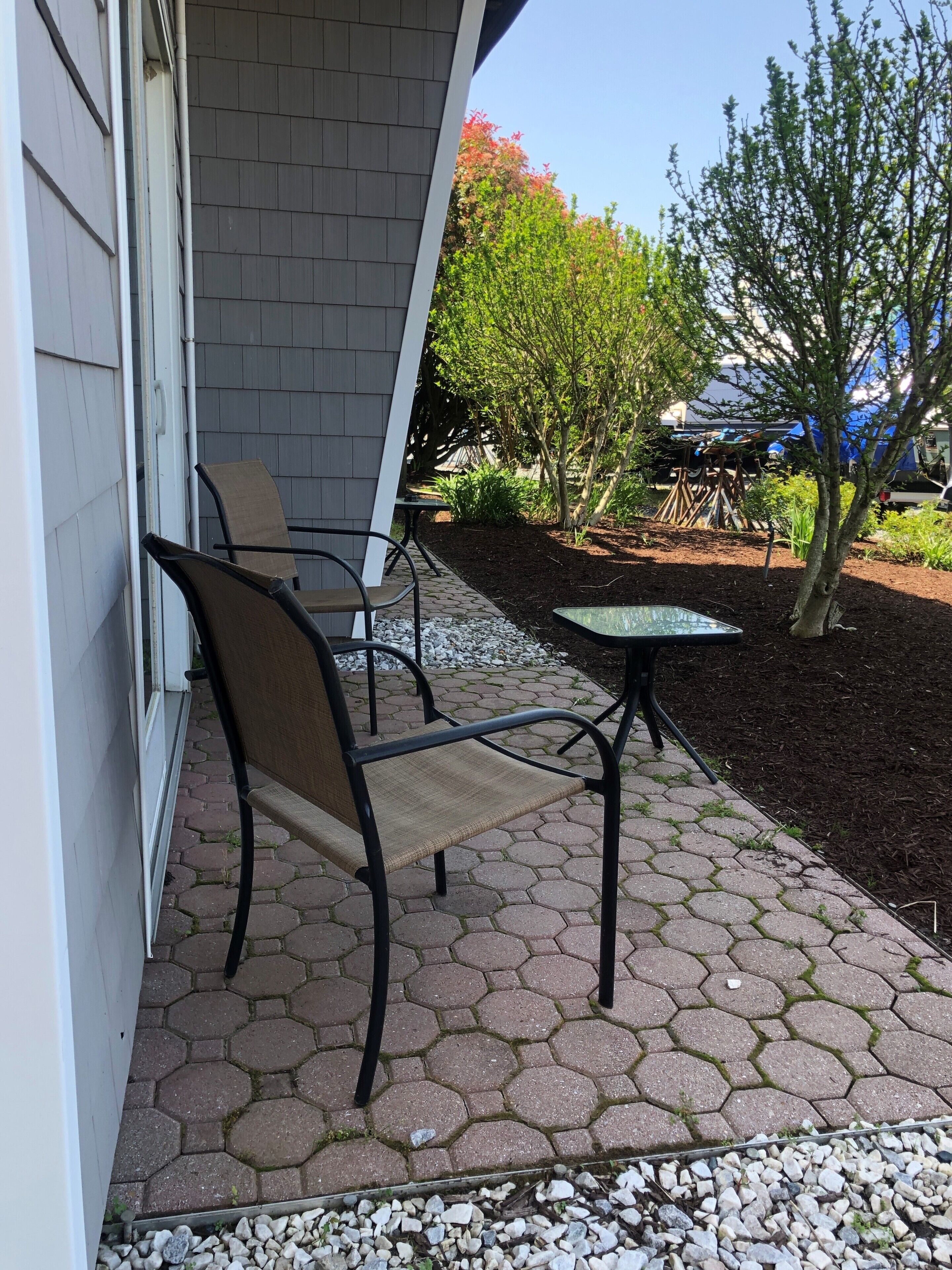 An outdoor patio area with hexagonal tiles, a metal chair, a small table, and a garden with various plants and a tree.