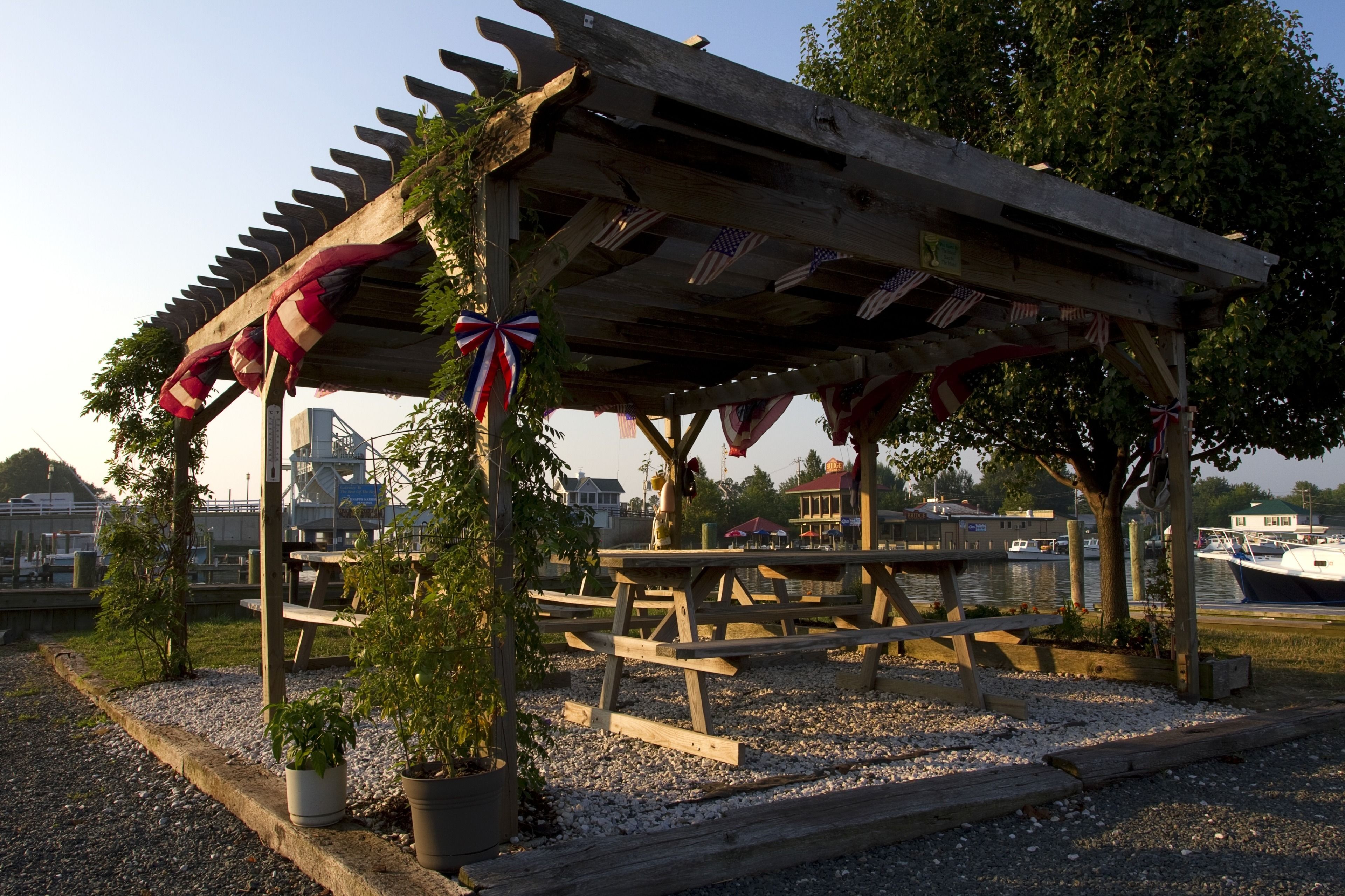 A wooden pavilion with picnic tables, surrounded by gravel and potted plants.