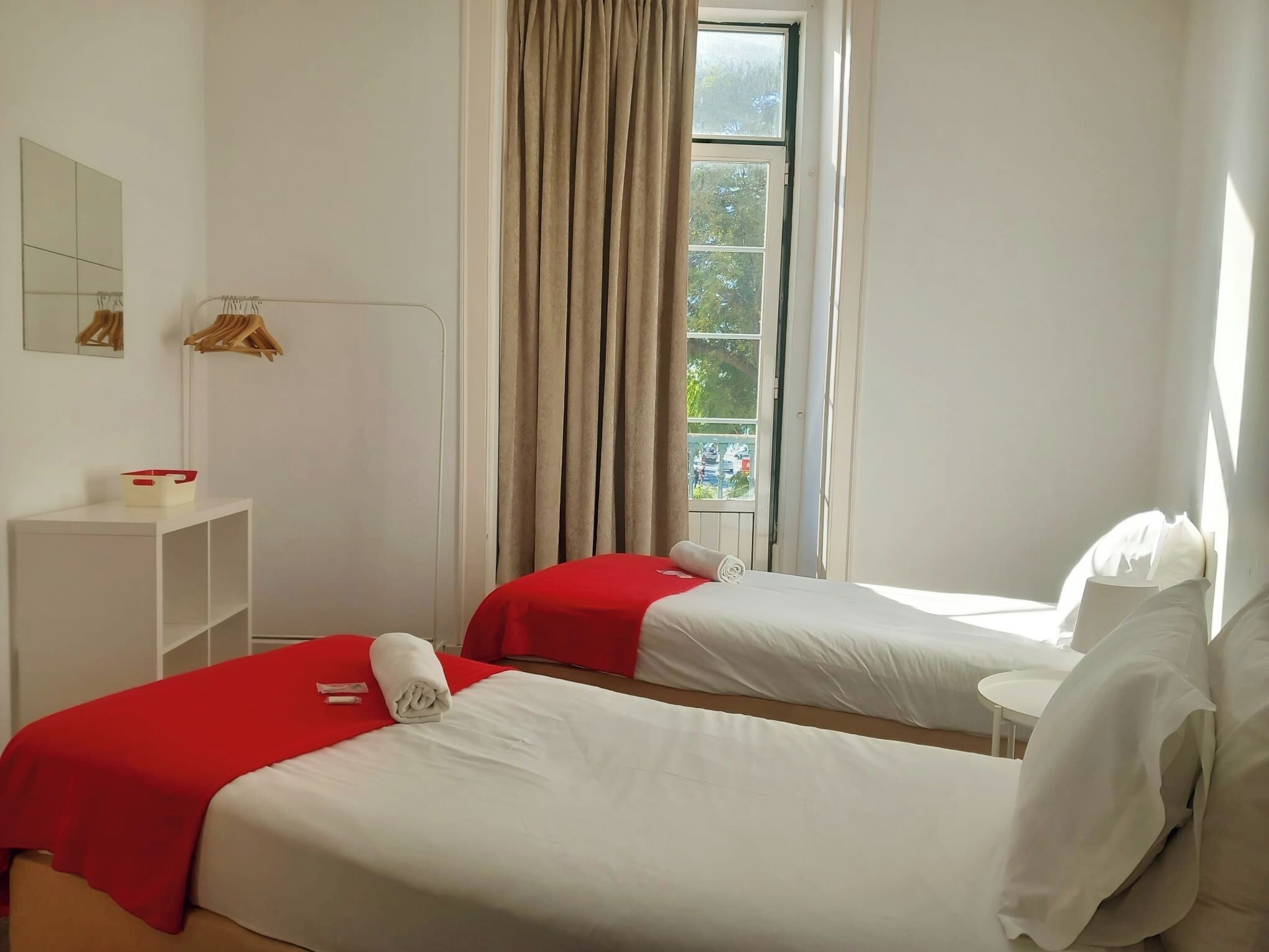 A hotel room with two beds, a window with curtains, a small table, and a mirror on the wall.