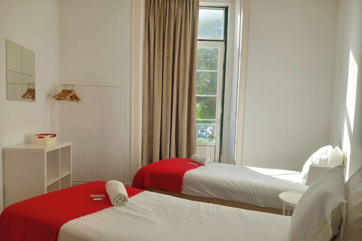 A hotel room with two beds, a window with curtains, a small table, and a mirror on the wall.