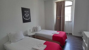 Habitación de hotel con dos camas individuales, una mesita, una silla, un ventanal con cortinas y un cartel en la pared.