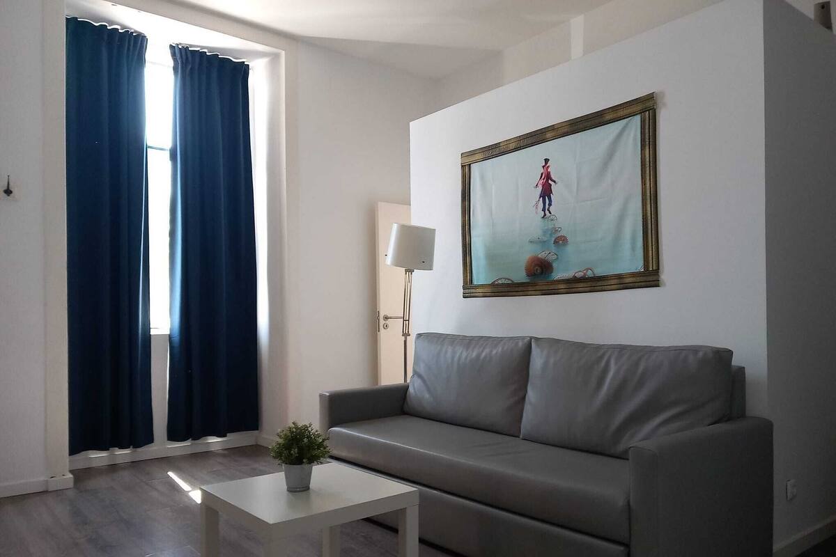 A modern living room with a grey sofa, a white coffee table, and a potted plant. A large framed picture hangs on the wall.