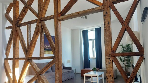Una sala de estar moderna con vigas de madera a la vista, un sofá gris, una mesa de centro blanca y vistas a un paisaje urbano a través de una ventana.