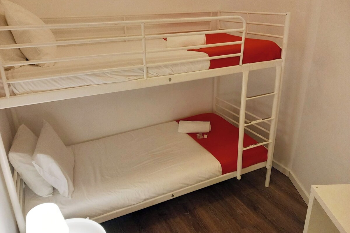A bunk bed with white and red bedding, a wooden floor, and a white desk.
