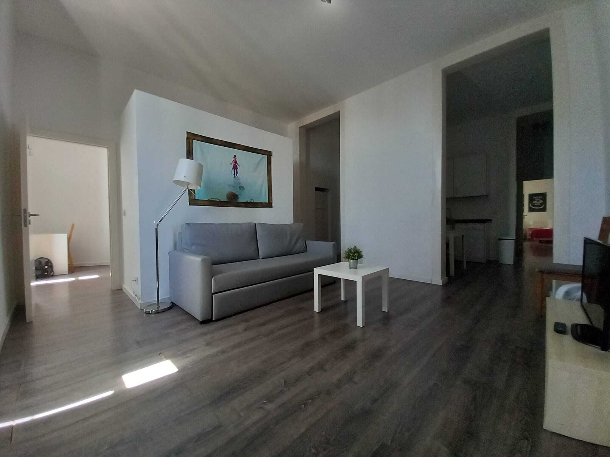 Una sala de estar moderna con un sofá gris, una mesita blanca pequeña y una lámpara de pie. Hay un cuadro en la pared y una puerta que da a otra habitación.