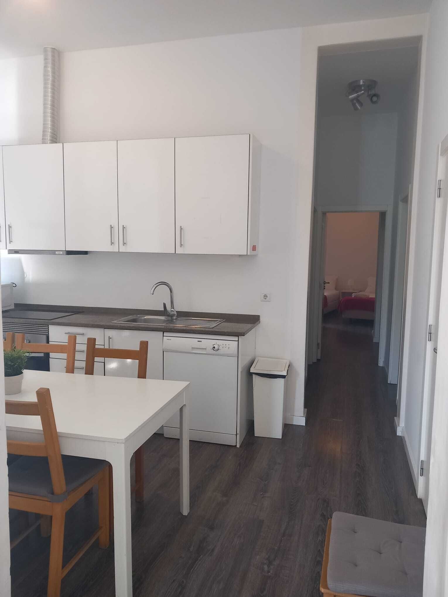 Cocina compacta con armarios blancos, lavavajillas y fregadero. Hay una mesa de comedor con sillas y un taburete pequeño. Se ve un cubo de basura.