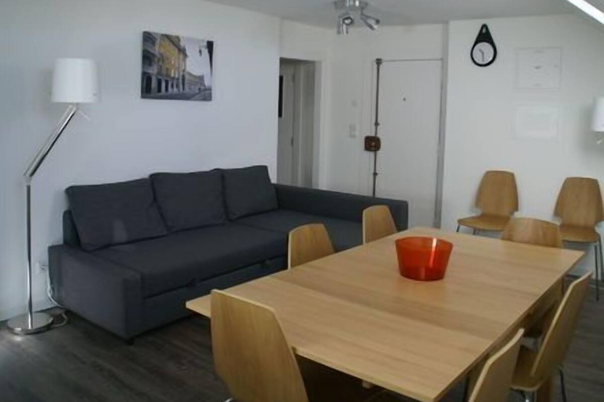 A modern living room with a dark grey sofa, a wooden dining table, and six wooden chairs. There is a floor lamp and a painting on the wall.