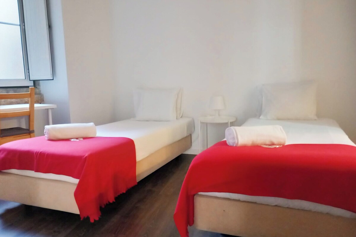 Two single beds with red blankets, white pillows, and a small white table with a lamp.