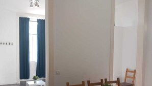 Un comedor con una mesa blanca y sillas de madera, una mesita blanca con una planta, y una ventana con cortinas azules.