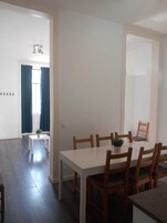 Un comedor con una mesa blanca y sillas de madera, una mesita blanca con una planta, y una ventana con cortinas azules.