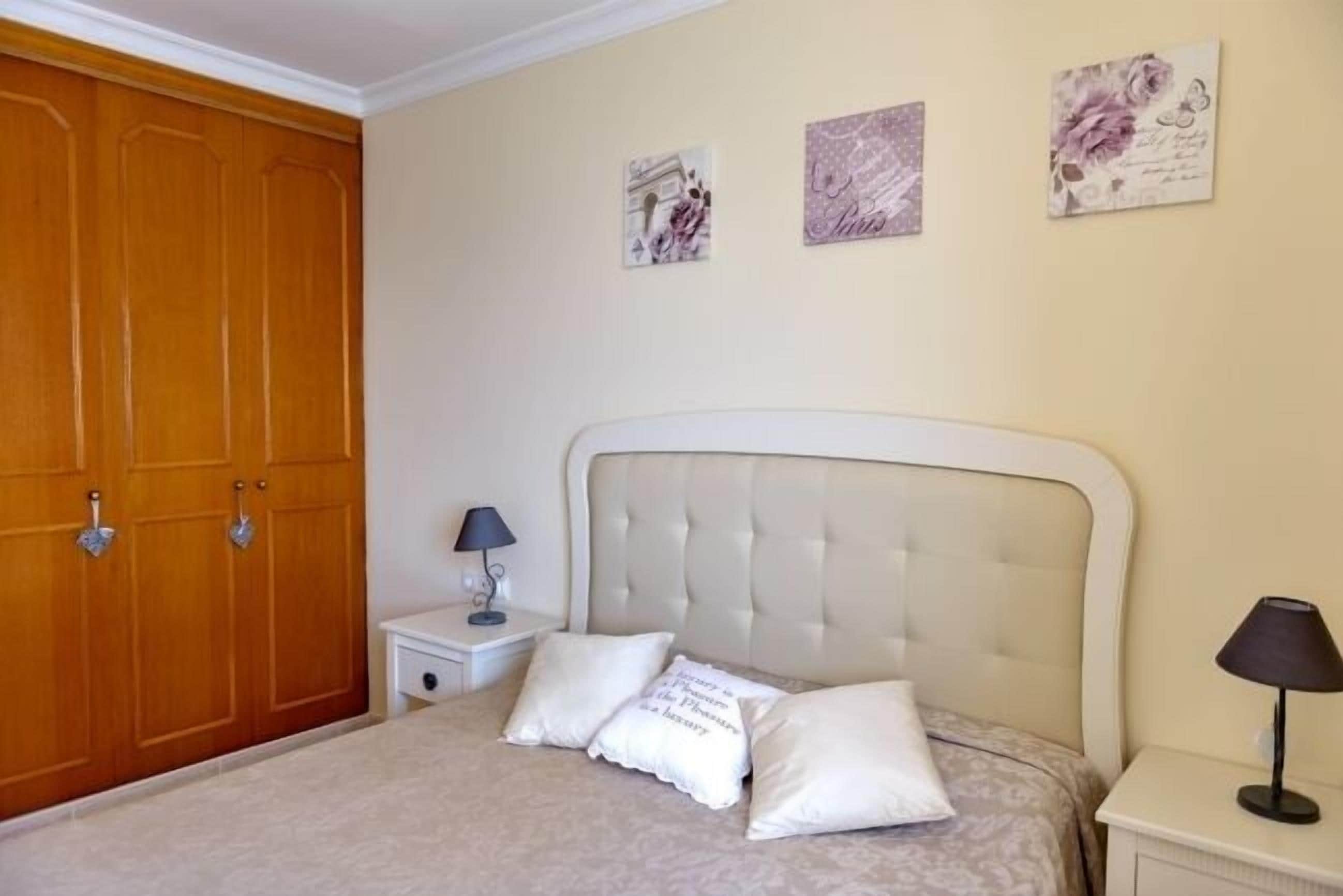 A bedroom with a large wooden wardrobe, a bed with a tufted headboard, two bedside tables with lamps, and three framed pictures on the wall.