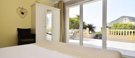 A bedroom with a large bed, a chair, a wardrobe, and a sliding glass door leading to a balcony with a view of a mountain.