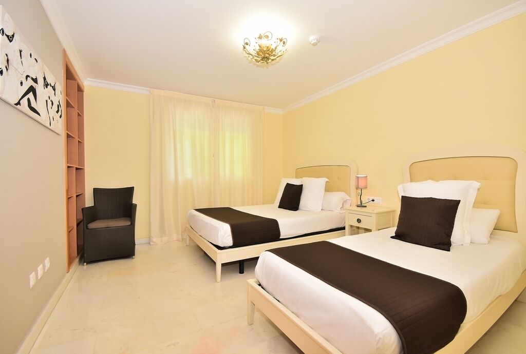 Una habitación de hotel con dos camas, una silla, una mesita de noche y un reloj en la pared.