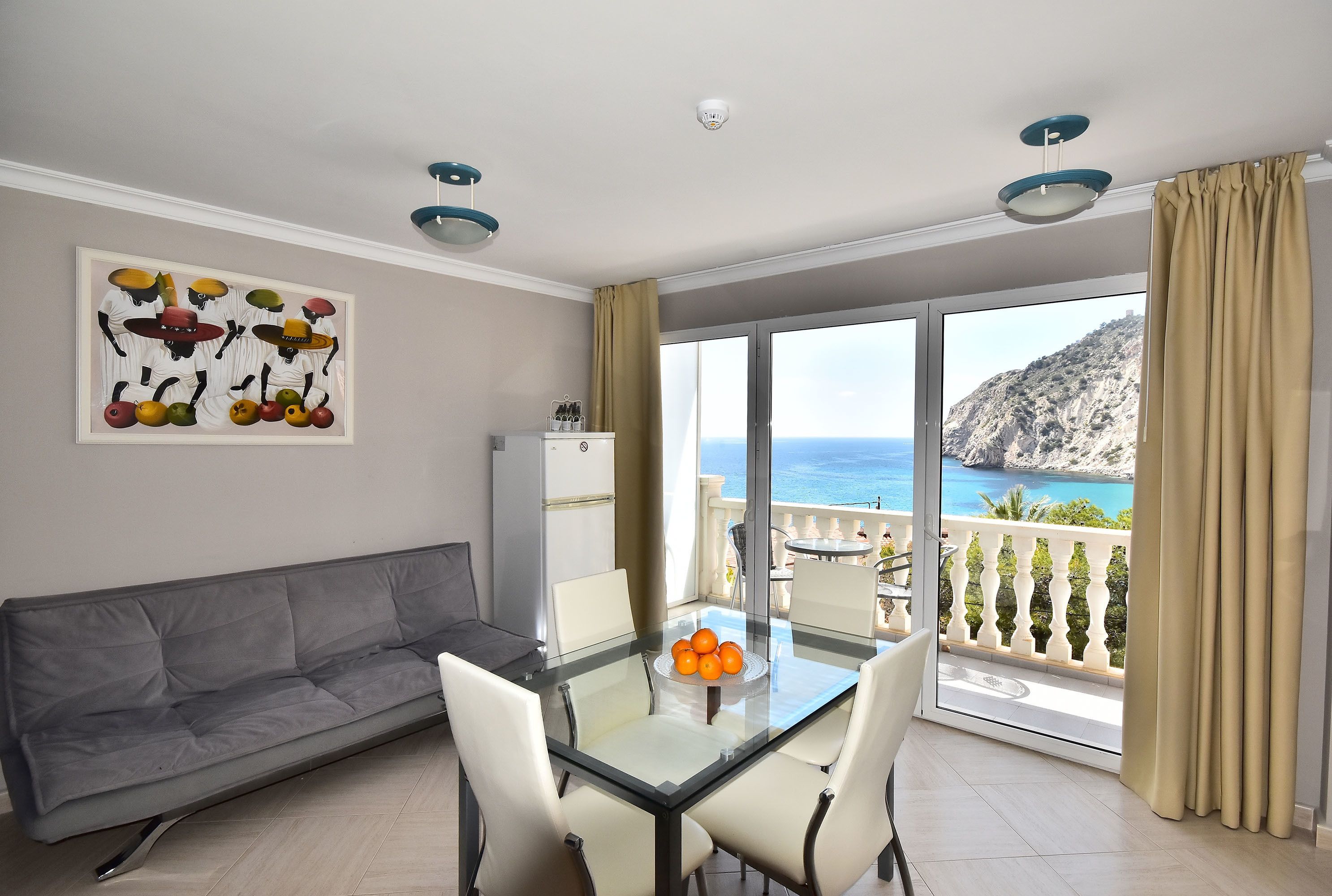 Un salon moderne avec un canapé gris, une table à manger en verre entourée de chaises, un réfrigérateur blanc et une grande fenêtre offrant une vue sur la mer et une montagne.
