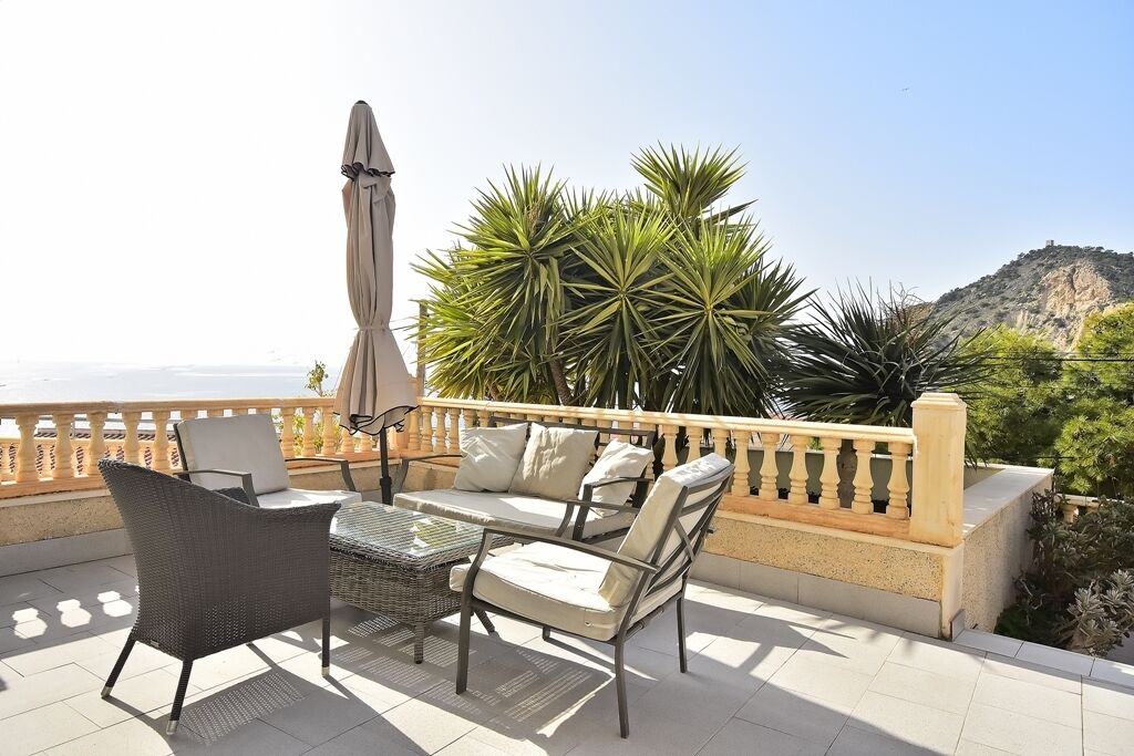 Une terrasse meublée avec des meubles en osier, une table et un parasol. On y trouve des plantes et, au loin, une vue sur une colline.
