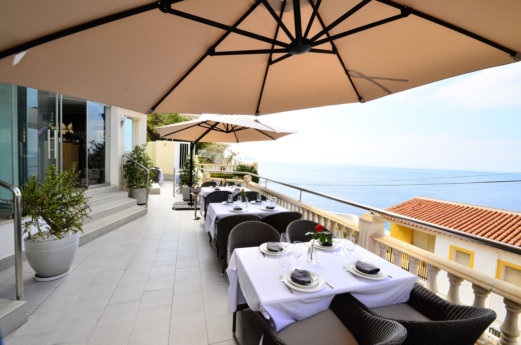 An outdoor dining area with tables and chairs, a large umbrella, and a view of the sea.