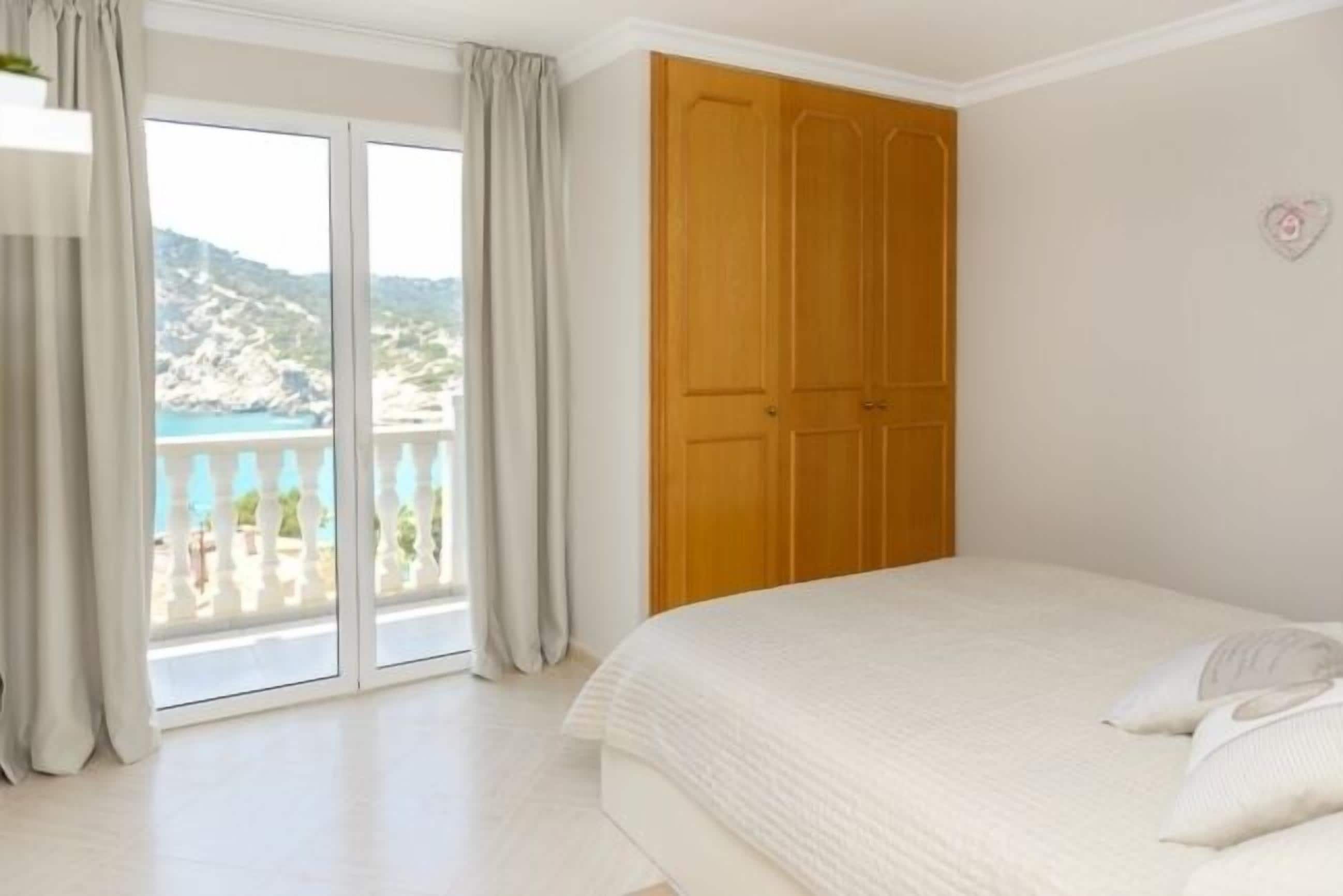 A bedroom with a large bed, a wooden wardrobe, and a balcony with a view of the sea.