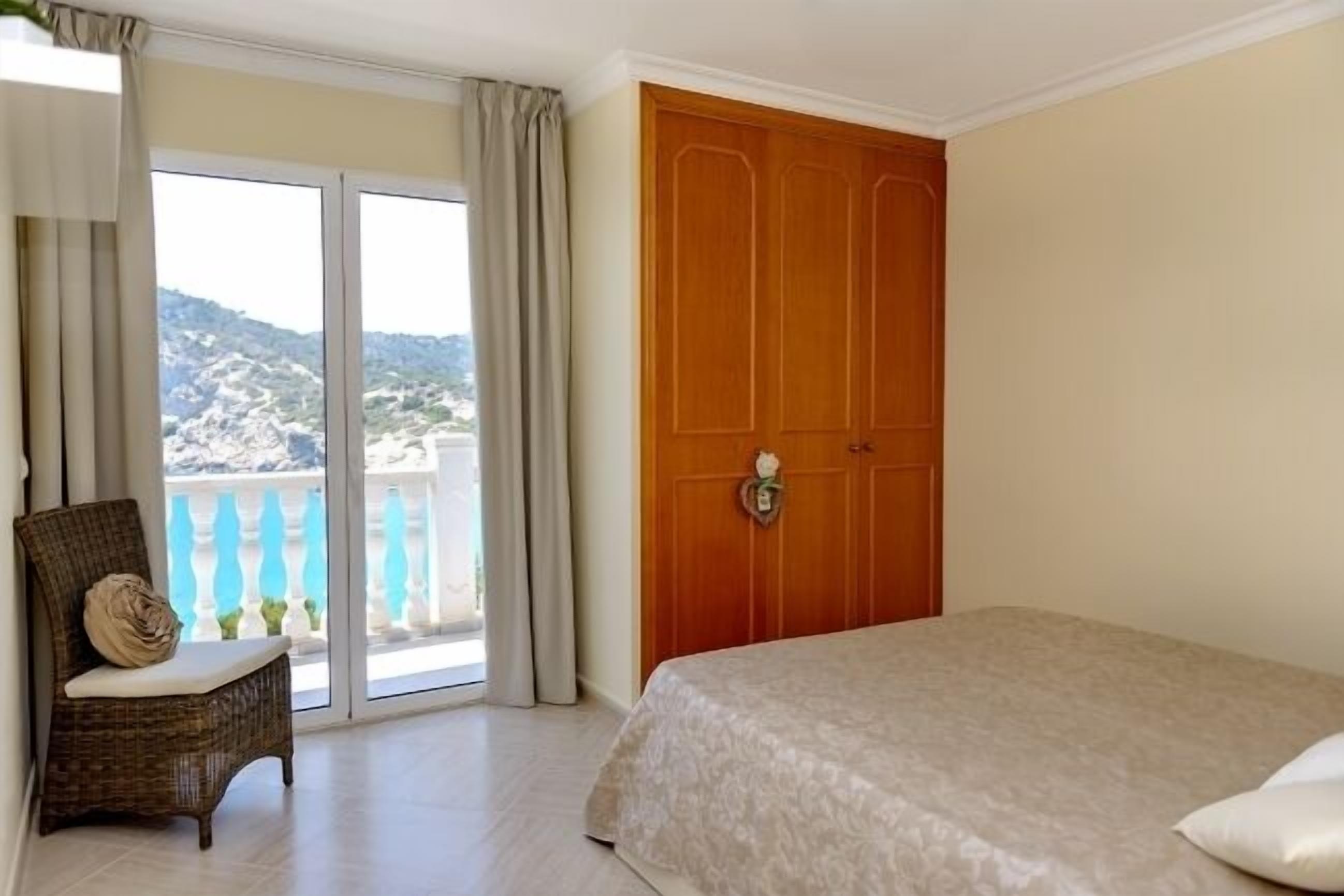 Um quarto com uma cama, uma cadeira de vime, uma porta de madeira e uma varanda com vista para as colinas.